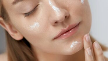 Foto aproximada do rosto de uma mulher, com os olhos fechados. Ela está passando um hidratante na pele, importante passo para manter a integridade da barreira cutânea.