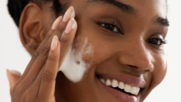 Imagem fechada no rosto de uma mulher. Ela tem a pele negra, está sorrindo e, com uma das mãos, passando uma espuma de limpeza facial na bochecha, um cuidado importante com o microbioma da pele.