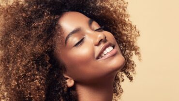 Imagem fechada no rosto de uma mulher negra. Ele está levemente inclinado para cima. Ela está com os olhos fechados e um sorriso. Sua pele está radiante e expressando plenitude.