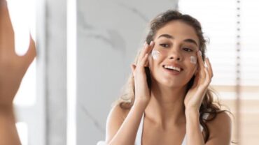 Imagem do rosto de uma mulher se olhando no espelho e aplicando produtos de skincare na face. Ela está sorrindo e com as duas mãos logo acima das bochechas, que estão cobertas de creme hidratante.