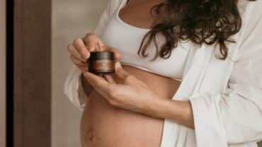 Imagem do tronco de uma mulher grávida. Ela está com um top e uma camisa, ambos branco, e segura um creme hidratante para o rosto com as duas mãos, dando a entender que iniciará sua rotina de skincare para gestantes.