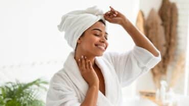 Mulher com os cabelos enrolados em uma toalha. Ela toca delicadamente a pele enquanto faz do skincare um ritual de conexão