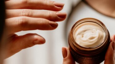 Imagem de duas mãos. A mão direita está segurando um pequeno pote de creme hidratante, enquanto a esquerda está indo em direção ao pote para tocar no produto.