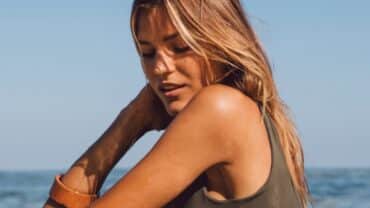 Imagem de uma mulher, com o mar ao fundo. A mulher é loira, com a pele bronzeada e veste uma regata marrom. Ela está olhando para si mesma, como se estivesse cuidando da pele no verão.