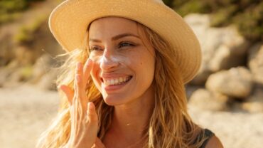 Imagem do rosto de uma mulher na praia. Ela está usando um chapéu de palha e passando protetor solar no rosto, duas medidas de fotoprotecao. Ela é loira, tem cabelos longos e está sorrindo.