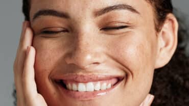 Imagem de uma mulher sorrindo, com os olhos fechados e uma das mãos no rosto. Ela tem cabelos escuros e cacheados presos e a pele possui sardas, um tipo de manchas de sol.
