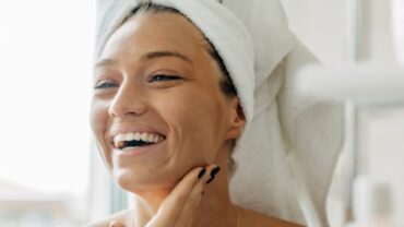Imagem fechada no rosto de uma mulher. Ela tem a pele clara, está com uma toalha de banho branca enrolada nos cabelos e parece estar se olhando no espelho. Está sorrindo e com uma das mãos próximo ao queixo.