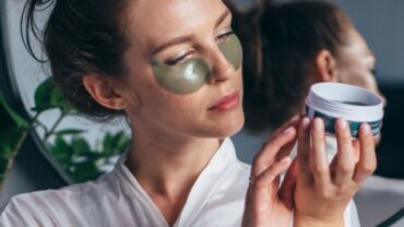 Imagem de umamulher branca, com os cabelos presos. Ela aplicou uma máscara facial verde abaixo dos olhos e esta lendo o rótulo do cosmético utilizado para avaliar se há ingredientes nocivos na formulação.