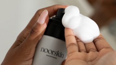 Imagem fechada da espuma de limpeza facial Light Velvet, da Noorskin. Na foto aparecem parte de duas mãos. Uma está apertando o frasco e a outra está segurando a espuma que sai dele.