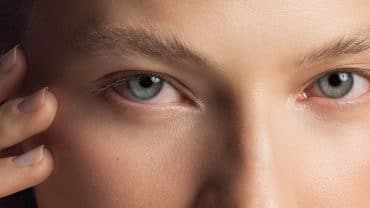 Imagem fechada nos olhos de uma mulher. Ela tem a pele e os olhos claros e está segurando o rosto com as mãos, próximo aos olhos, como se estivesse puxando a pele para trás.