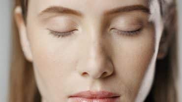 Imagem fechada no rosto de uma mulher. Ela tem a pele e os cabelos claros e está com os olhos fechados, como se estivesse em um momento de atenção plena, uma prática importante para equilibrar a conexão mente e pele.