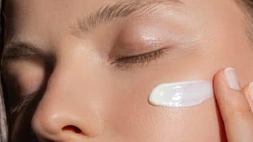 Imagem fechada na parte superior do rosto de uma mulher. Ela tem a pele clara, está de olhos fechados e passa protetor solar na face com os dedos.