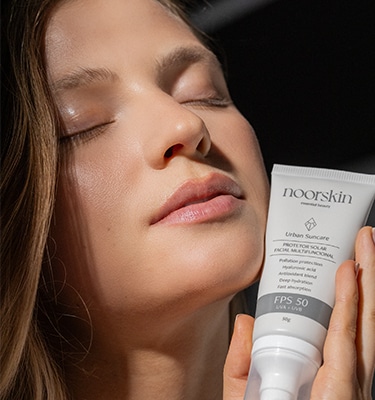 Imagem focada no rosto de uma mulher e no protetor solar facial da Noorskin, o Urban Suncare. A mulher tem a pele clara e está de olhos fechados, segurando o produto em uma das mãos.