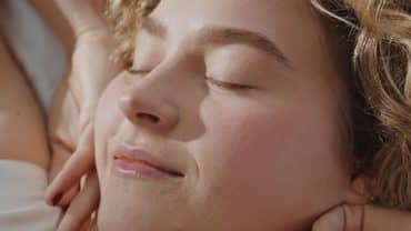 Imagem fechada no rosto de uma mulher. Ela tem a pele e os cabelos claros, está de olhos fechados e com as mãos nas laterais do roso, como se estivessem acabando de acordar do seu sono da beleza.
