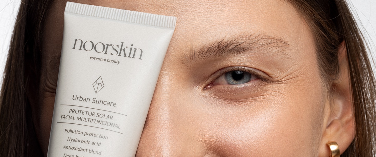 Imagem fechada na região dos olhos de uma mulher. Ela tem a pele e os olhos claros e está com o frasco do protetor solar facial da Noorskin colado ao rosto.